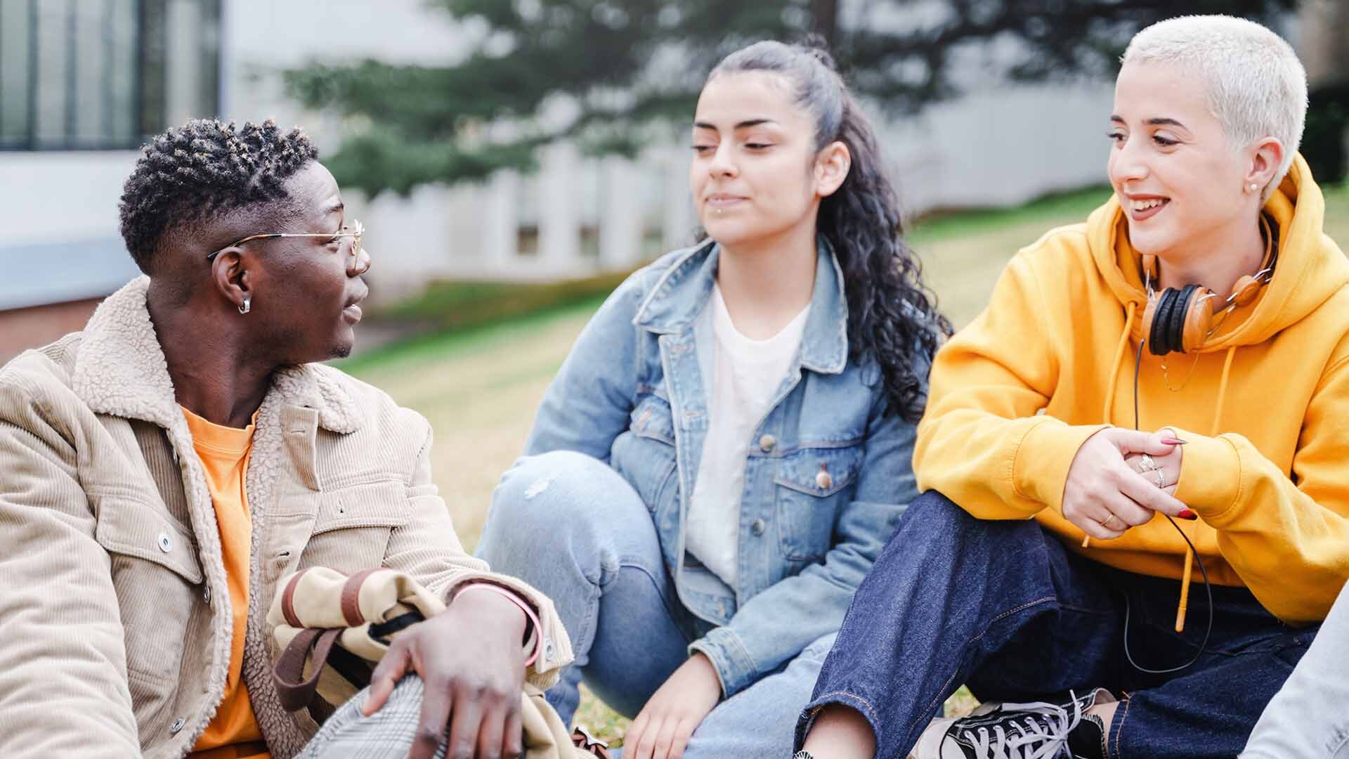 Chico y dos chicas en la calle sentados en el césped de una universidad conversando