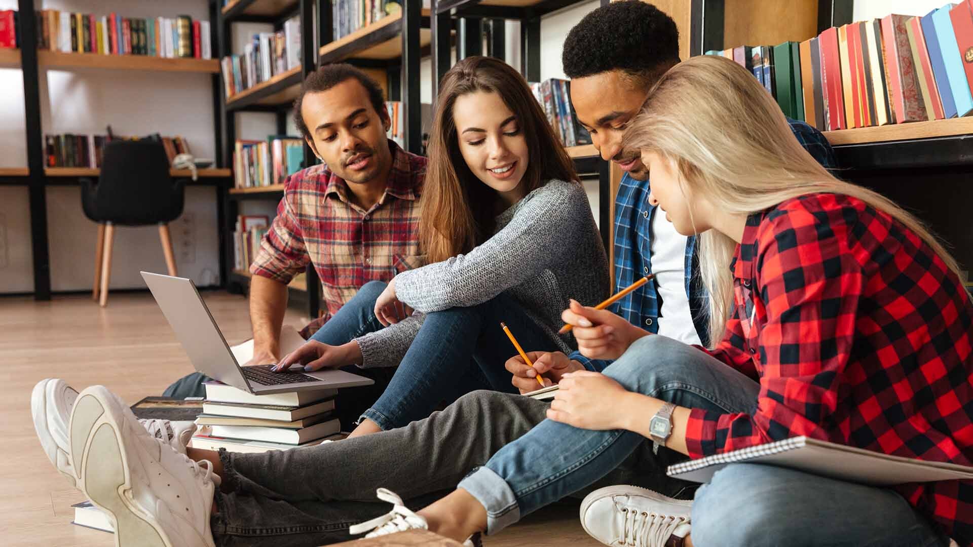 Dos chicos y chicas de rasgos fenotípicos diferentes sentados en el suelo en una biblioteca y apoyados en una estantería. Miran con interés y diversión uno de los libros sostenidos por uno de los chicos.