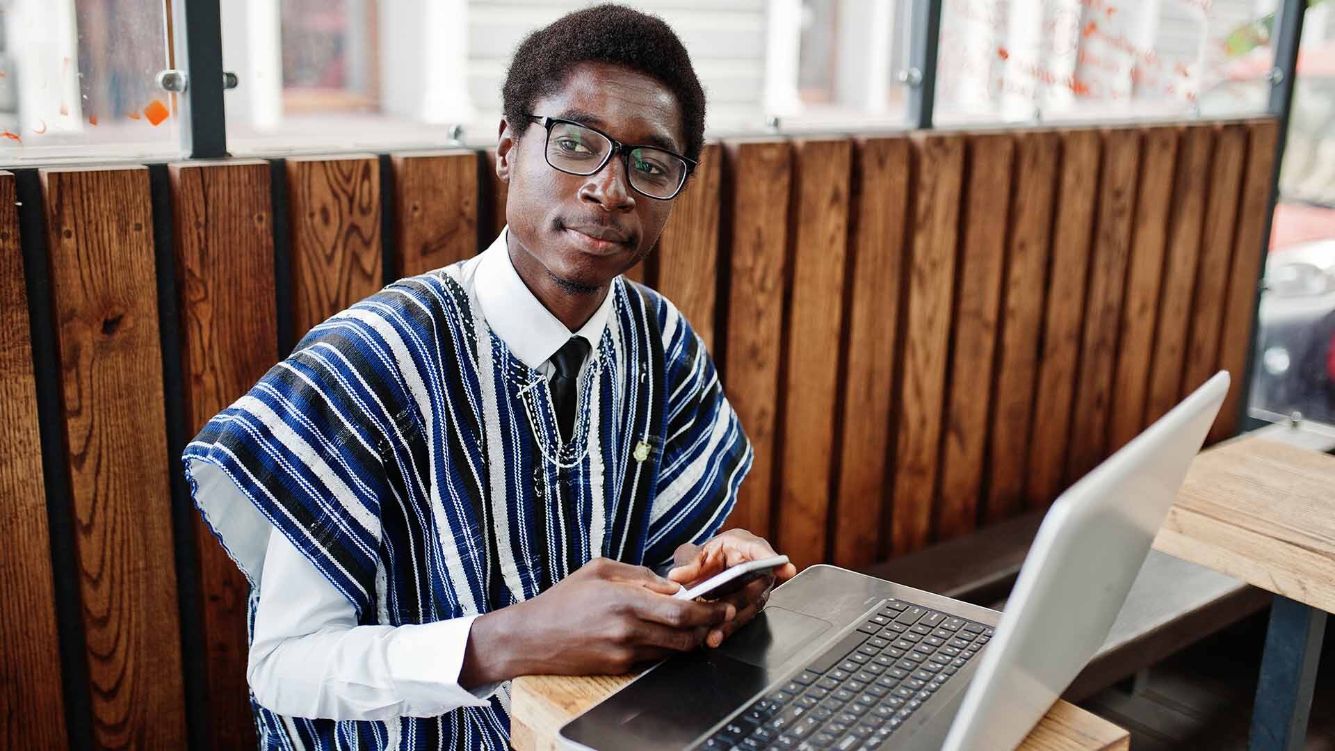 Hombre con vestimenta y rasgos fenotípicos africanos en una mesa sentado con su ordenado y móvil trabajando.